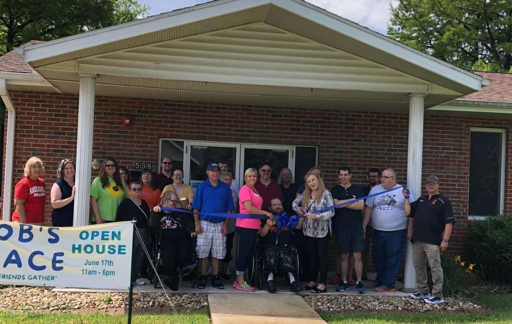 brick building with white triangular awing over entryway, people standing behind a large long blue ribbon